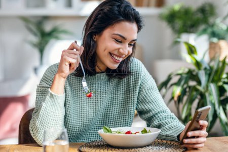 Téléchargez les photos : Tourné de belle jeune femme mangeant un bol de salade tout en utilisant son téléphone portable dans le salon à la maison. - en image libre de droit