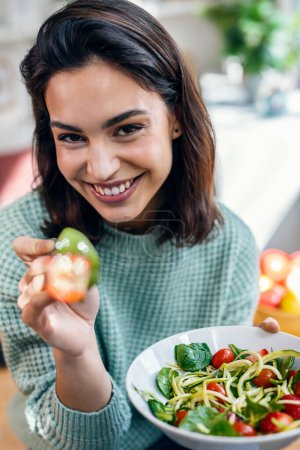 Photo for Shot of beautiful smiling woman eating healthy salad while sitting on the table at home. - Royalty Free Image