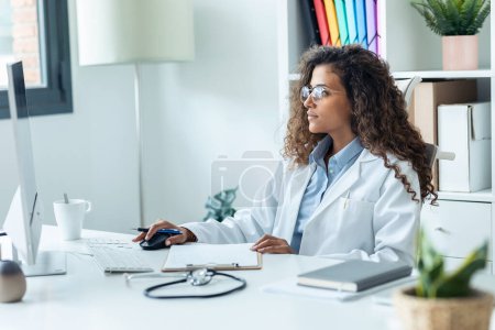 Foto de Fotografía de una hermosa joven doctora haciendo videocall con paciente en computadora mientras toma algunas notas en la consulta del hospital - Imagen libre de derechos