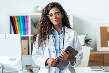 Porträt der schönen jungen Ärztin, die ein digitales Tablet in der Hand hält, während sie in die Kamera blickt, die in der Krankenhaussprechstunde steht