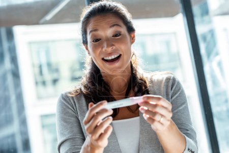 Shot of happy woman celebrating while looking at predictor finding out she is pregnant at home