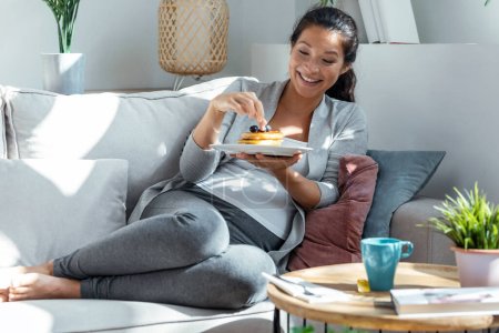 Photo for Shot of beautiful happy pregnant woman in panties eating pancakes with red fruits lying on sofa at home - Royalty Free Image