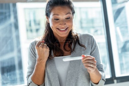 Téléchargez les photos : Prise de vue de femme heureuse célébrant tout en regardant prédicteur découvrir qu'elle est enceinte à la maison - en image libre de droit