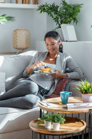 Téléchargez les photos : Tourné de belle femme enceinte heureuse en culotte manger des crêpes avec des fruits rouges couché sur le canapé à la maison - en image libre de droit