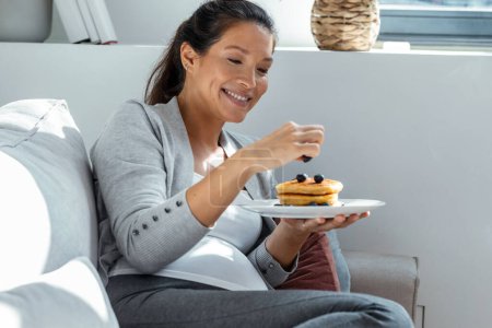 Téléchargez les photos : Tourné de belle femme enceinte heureuse en culotte manger des crêpes avec des fruits rouges couché sur le canapé à la maison - en image libre de droit