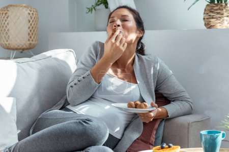 Aufnahme der schönen glücklichen schwangeren Frau in Höschen, die zu Hause auf dem Sofa liegend Schokoladentrüffel isst