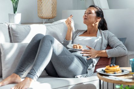 Téléchargez les photos : Tourné de belle femme enceinte heureuse en culotte manger des truffes au chocolat couché sur le canapé à la maison - en image libre de droit