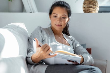 Téléchargez les photos : Prise de vue de belle femme enceinte relaxante lisant un livre tout en mangeant des crêpes aux fruits rouges couché sur le canapé à la maison - en image libre de droit