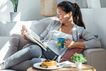Foto de Tiro de hermosa mujer embarazada relajante leyendo un libro mientras que la taza de té tumbado en el sofá en casa - Imagen libre de derechos