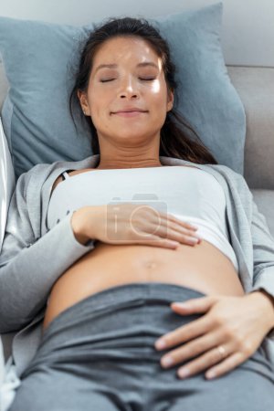 Foto de Shot de hermosa mujer embarazada tocando su vientre mientras se relaja tumbado en el sofá en casa - Imagen libre de derechos