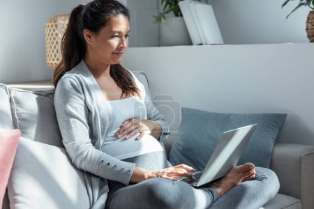 Téléchargez les photos : Prise de vue de belle femme enceinte travaillant avec un ordinateur portable assis sur le canapé dans le salon à la maison. - en image libre de droit