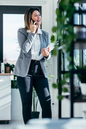 Téléchargez les photos : Prise de vue de belle femme d'affaires élégante travaillant tout en parlant avec smartphone debout à côté de la fenêtre dans un bureau moderne - en image libre de droit