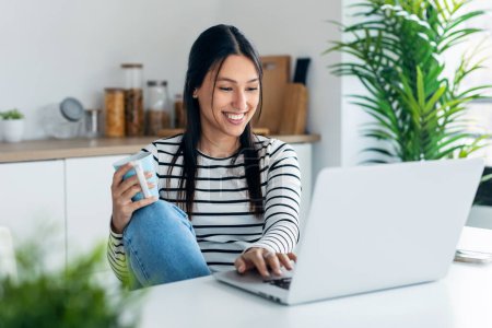 Foto de Fotografía de la joven sonriente haciendo videollamada con el ordenador portátil mientras sostiene una taza de café en la cocina en casa. - Imagen libre de derechos