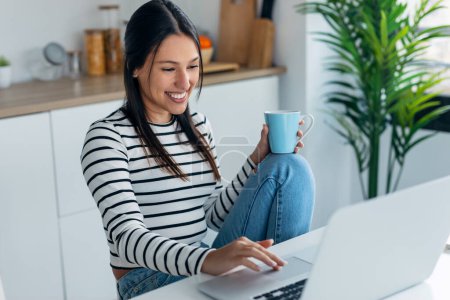 Téléchargez les photos : Prise de vue d'une jeune femme souriante faisant un appel vidéo avec un ordinateur portable tout en tenant une tasse de café dans la cuisine à la maison. - en image libre de droit