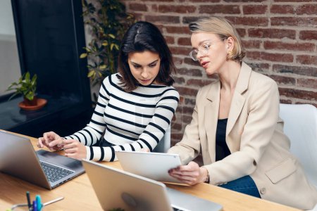 Photo for Shot of two beautiful designer women working with laptop while choosing materials in digital tablet in the modern office. - Royalty Free Image