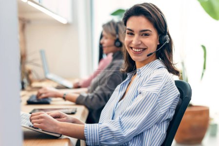 Photo for Shot of group of female operator talking with wireless headset while searching information in laptop in the office - Royalty Free Image