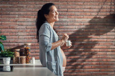 Téléchargez les photos : Tourné de belle femme enceinte gaie avec bretelles manger un yaourt dans la cuisine à la maison - en image libre de droit