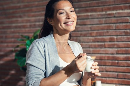Téléchargez les photos : Tourné de belle femme enceinte gaie avec bretelles manger un yaourt dans la cuisine à la maison - en image libre de droit