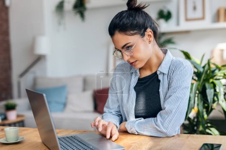 Téléchargez les photos : Tourné de belle femme entrepreneure attrayante travaillant avec un ordinateur portable tout en buvant du café dans le salon à la maison. - en image libre de droit