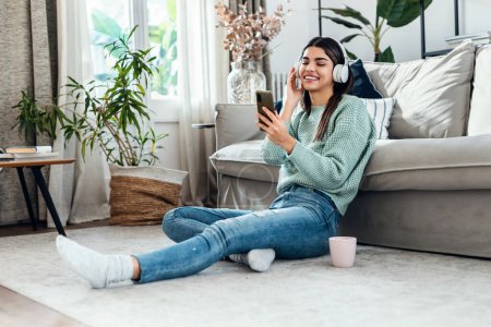 Téléchargez les photos : Prise de vue d'une jeune femme motivée écoutant de la musique avec un smartphone tout en dansant assise sur le sol à la maison. - en image libre de droit