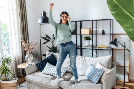Foto de Foto de mujer divertida escuchando música con teléfono inteligente mientras baila en el sofá en la sala de estar en casa - Imagen libre de derechos