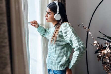 Foto de Foto de dulce mujer joven escuchando música con auriculares mientras mira hacia adelante a través de la ventana en casa - Imagen libre de derechos
