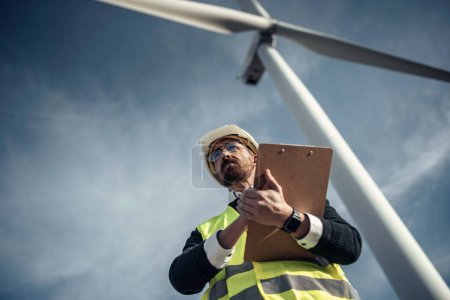 Téléchargez les photos : Prise de vue d'un ingénieur mâle mature en train de vérifier en prenant des notes sur le fonctionnement des éoliennes - en image libre de droit