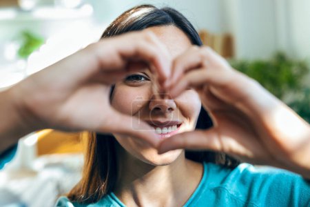 Photo for Portrait of romantic happy woman making heart shape with hands while smiling to camera at home - Royalty Free Image