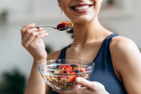 Foto de Foto de mujer atlética comiendo un tazón saludable de muesli con fruta en la cocina en casa - Imagen libre de derechos