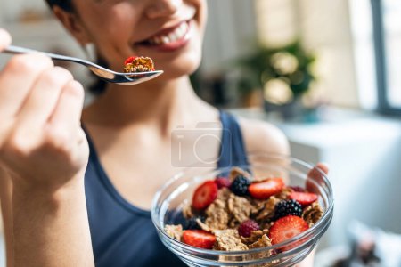 Téléchargez les photos : Tournage d'une femme sportive mangeant un bol sain de muesli avec des fruits dans la cuisine à la maison - en image libre de droit