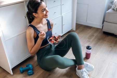 Téléchargez les photos : Prise de vue d'une femme sportive mangeant un bol sain de muesli avec des fruits assis sur le sol dans la cuisine à la maison - en image libre de droit