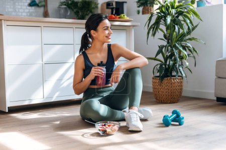 Foto de Tiro de mujer atlética bebiendo batido de proteína saludable mientras está sentado en el suelo en la cocina en casa - Imagen libre de derechos