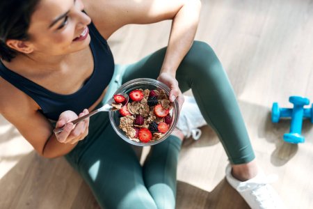 Aufnahme einer athletischen Frau beim Essen einer gesunden Schüssel Müsli mit Früchten, die zu Hause in der Küche auf dem Boden sitzt