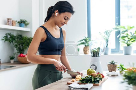 Aufnahme einer sportlichen Frau, die Obst und Gemüse schneidet, um einen Smoothie zuzubereiten, während sie zu Hause in der Küche mit Kopfhörern Musik hört
