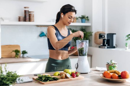 Shot of athletic woman preparing smothie with vegetables and fruits while listening music with earphones in the kitchen at home.
