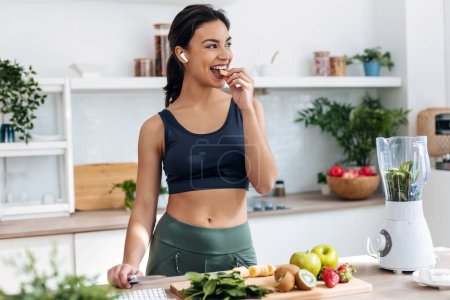 Sesión de mujer atlética cortando frutas y verduras para preparar un batido mientras escucha música con auriculares en la cocina en casa