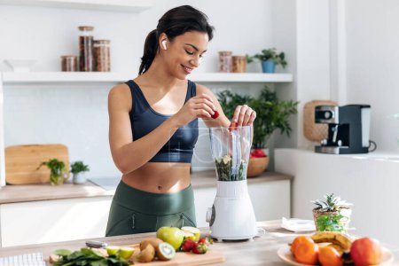 Foto de mujer atlética preparando smothie con verduras y frutas mientras escucha música con auriculares en la cocina en casa.