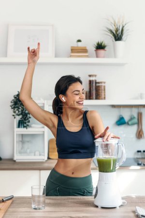 Photo for Shot of athletic woman preparing smothie while singing, dancing and listening music with earphones in the kitchen at home. - Royalty Free Image