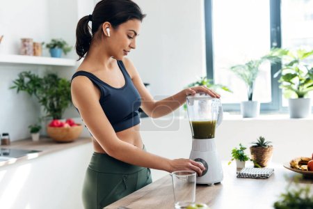 Athletic woman preparing smothie while listening music with earphones in the kitchen at home