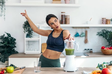 Photo for Shot of athletic woman preparing smothie while singing, dancing and listening music with earphones in the kitchen at home. - Royalty Free Image