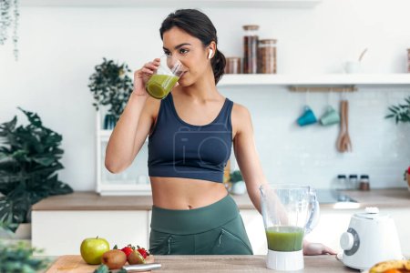 Photo for Shot of athletic smiling woman drinking smothie while listening music with earphones in the kitchen at home. - Royalty Free Image
