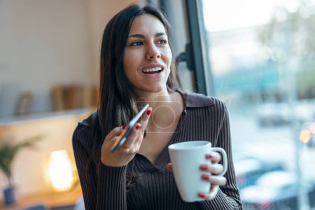 Téléchargez les photos : Prise de vue de jolie jeune femme utilisant son téléphone portable tout en buvant du café près de la fenêtre dans le salon à la maison - en image libre de droit