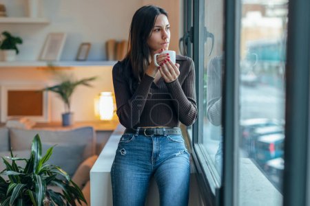 Téléchargez les photos : Tourné de belle femme réfléchie buvant une tasse de café tout en attendant avec impatience à côté de la fenêtre à la maison - en image libre de droit