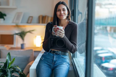 Téléchargez les photos : Prise de vue de belle femme réfléchie buvant une tasse de café tout en regardant la caméra à côté de la fenêtre à la maison - en image libre de droit