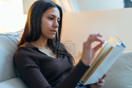 Photo for Shot of pretty young woman reading a book while sitting on sofa at home - Royalty Free Image