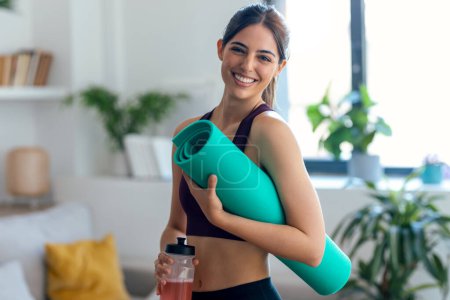 Téléchargez les photos : Prise de vue d'une femme sportive tenant un tapis de yoga tout en buvant une boisson protéinée en regardant la caméra à la maison - en image libre de droit