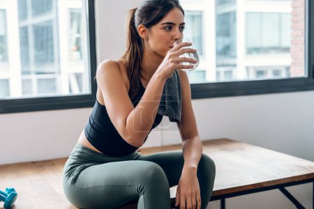Photo for Shot of sporty woman taking a break from exercising while drinking a glass of water sitting at home - Royalty Free Image