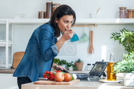 Foto de Tiro de hermosa mujer joven cocinando alimentos saludables y el uso de tabletas digitales para recetas en la cocina en casa. - Imagen libre de derechos