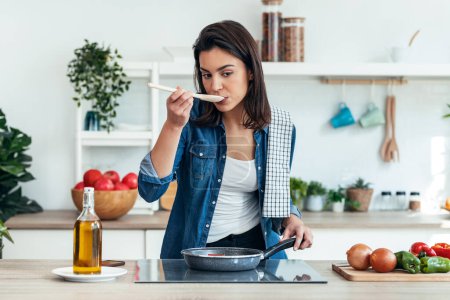 Photo for Shot of healthy young woman cooking and tasting food with wooden spoon in the kitchen at home - Royalty Free Image