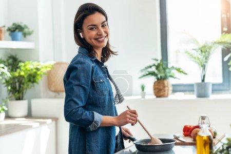 Foto de Foto de hermosa mujer sonriente cocinando verduras frescas mientras escucha música con auriculares en la cocina en casa - Imagen libre de derechos